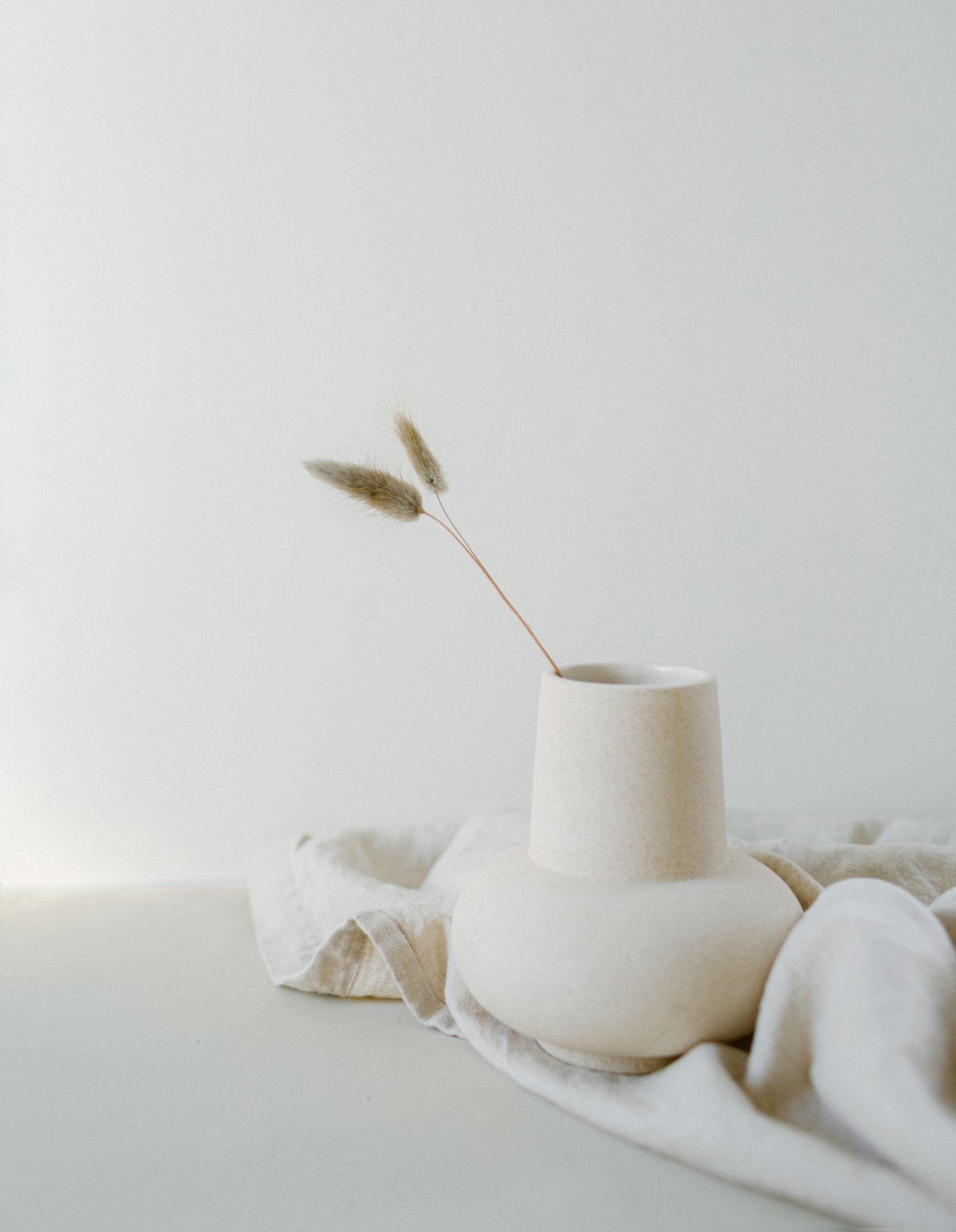 A single dried stem in a pale ceramic vase resting on soft linen