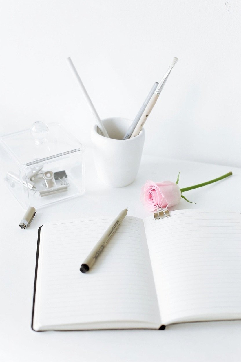 An open notebook, a pen and a single pale pink rose on a white desk